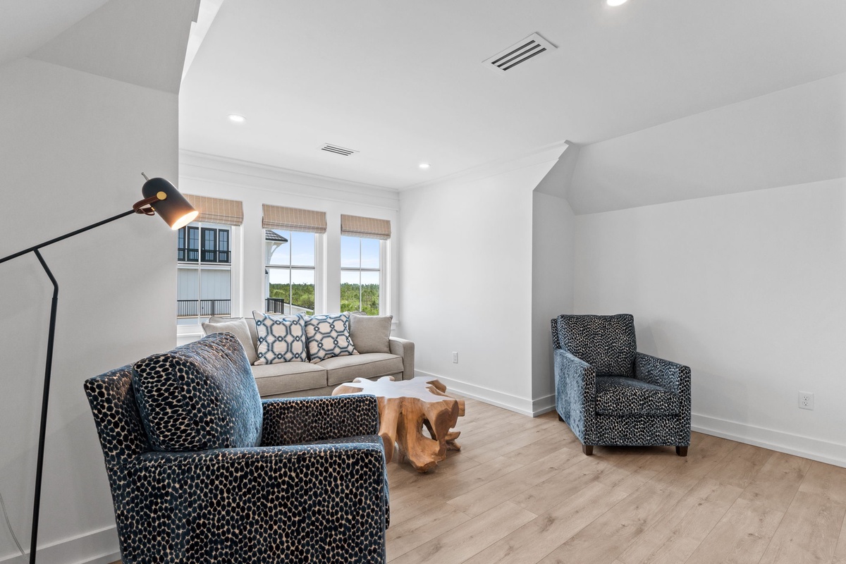 Relax in your bright, airy living room with plush seating, natural light streaming through bamboo shades, and serene views of greenery outside. The warm wood floors and modern furnishings create a welcoming retreat.