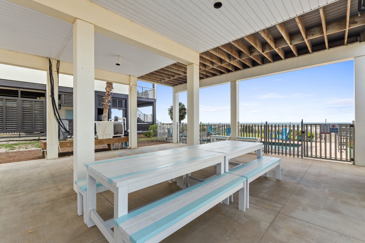 Covered patio area featuring whitewashed picnic tables with turquoise accents, overlooking water views, with nearby BBQ setup for outdoor dining.