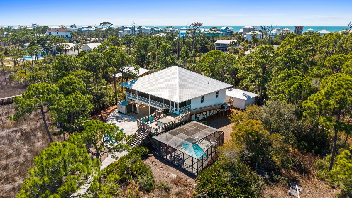 Elevated beachfront home nestled among towering pines with turquoise waters visible beyond the coastal community.