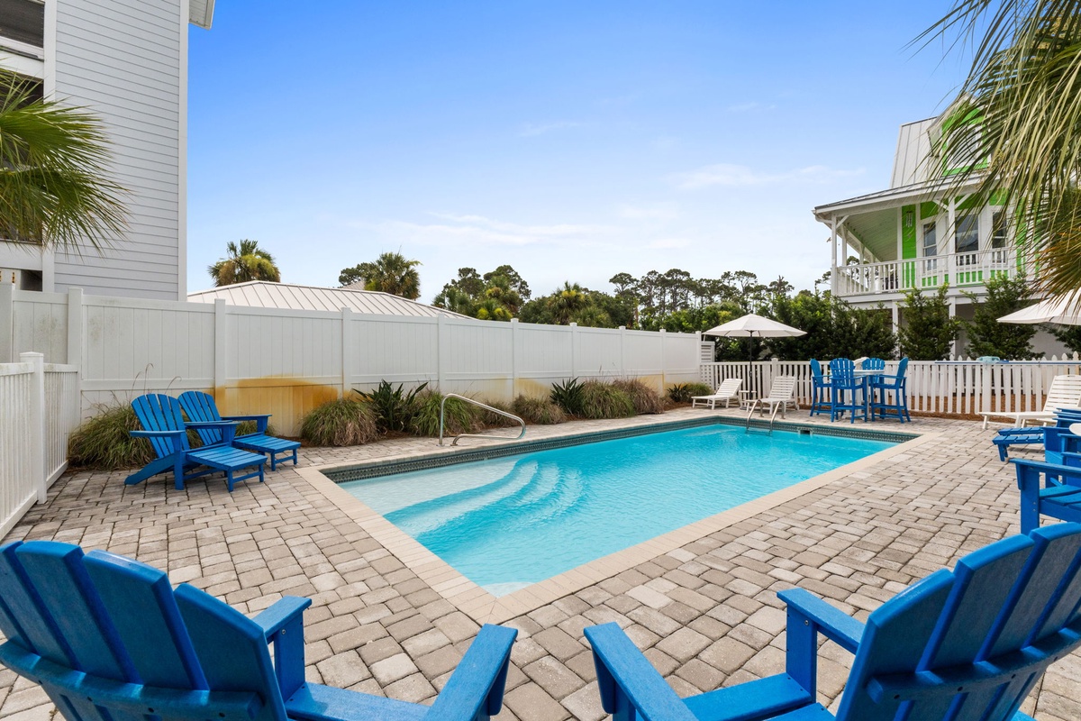 A coastal haven features an expansive private pool surrounded by vibrant blue lounge chairs on brick pavers, with tropical palms swaying overhead and neighboring homes framing this sunny retreat under clear skies.