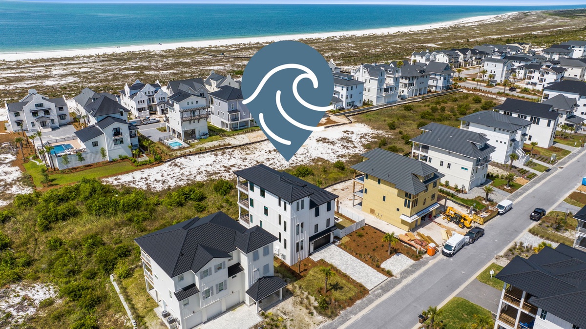 Aerial view of a coastal residential community with modern beach houses nestled between pristine white sand beaches and turquoise ocean waters. The neighborhood is part of a scenic beachfront area.