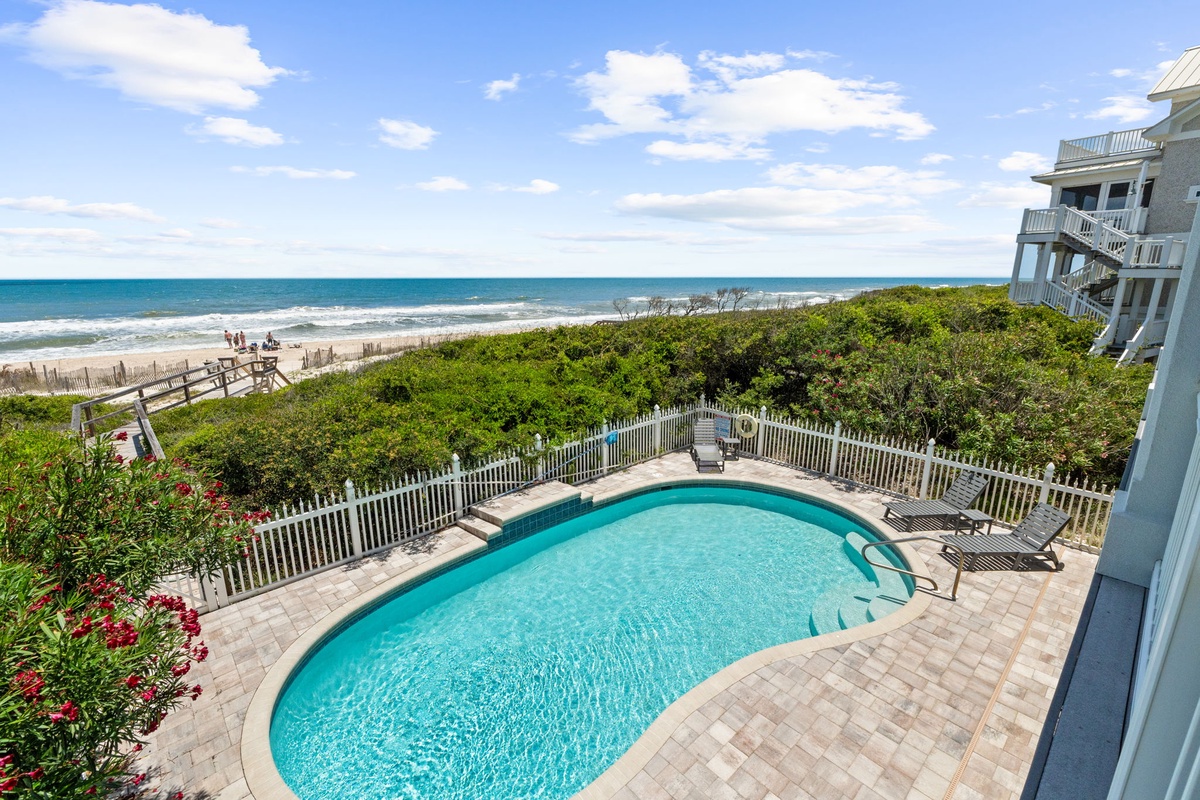 A beachfront property featuring a private pool, direct beach access, and sweeping ocean views framed by coastal greenery and a clear blue sky.
