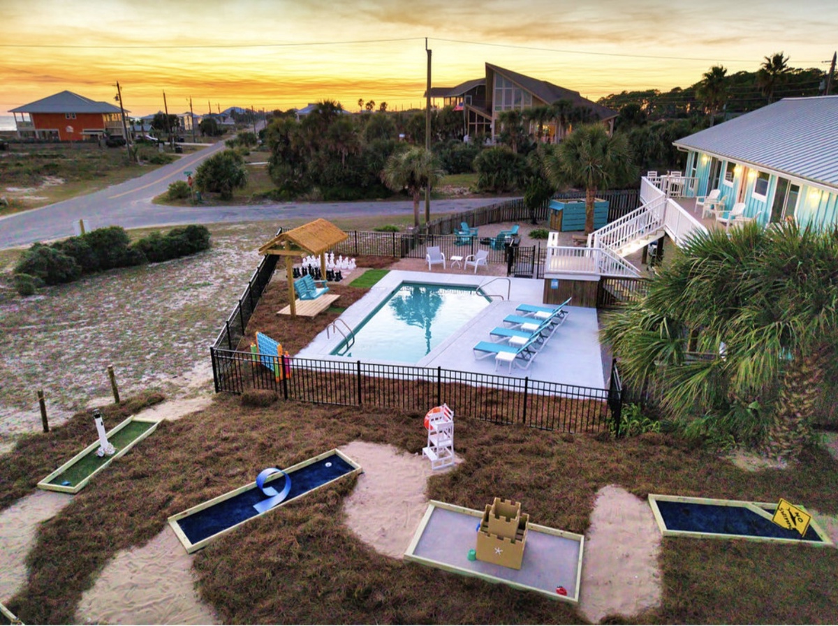 Aerial view of a coastal vacation rental featuring swimming pool, miniature golf course, and outdoor recreation areas surrounded by tropical landscape at sunset.
