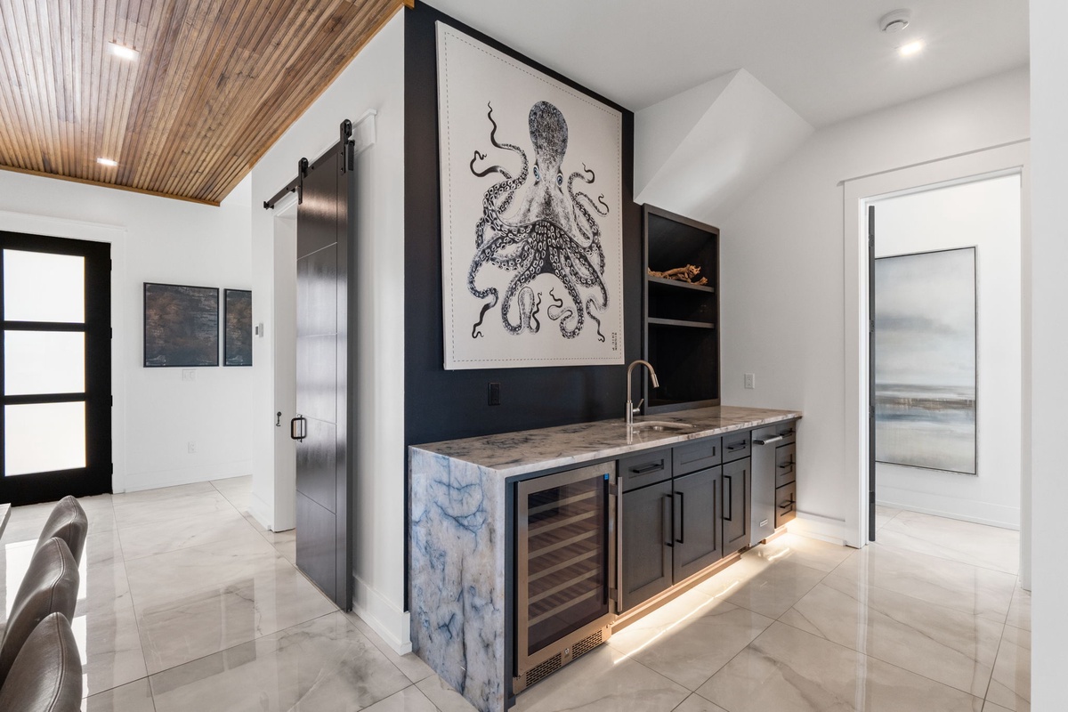Your stylish wet bar with wine fridge, sleek dark cabinetry, marble countertops, and striking octopus artwork creates the perfect spot to unwind with refreshments after a day of exploring.