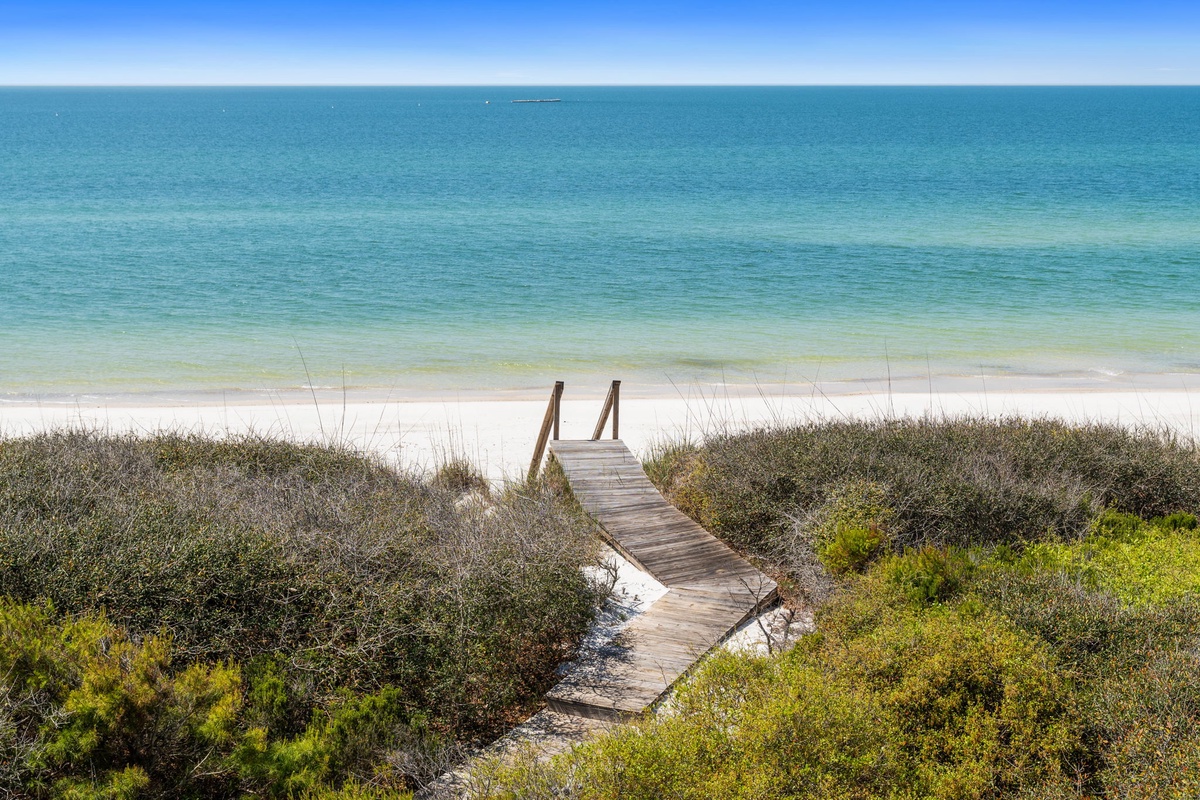 A wooden boardwalk leads through coastal dunes to pristine turquoise waters and white sand beach.
