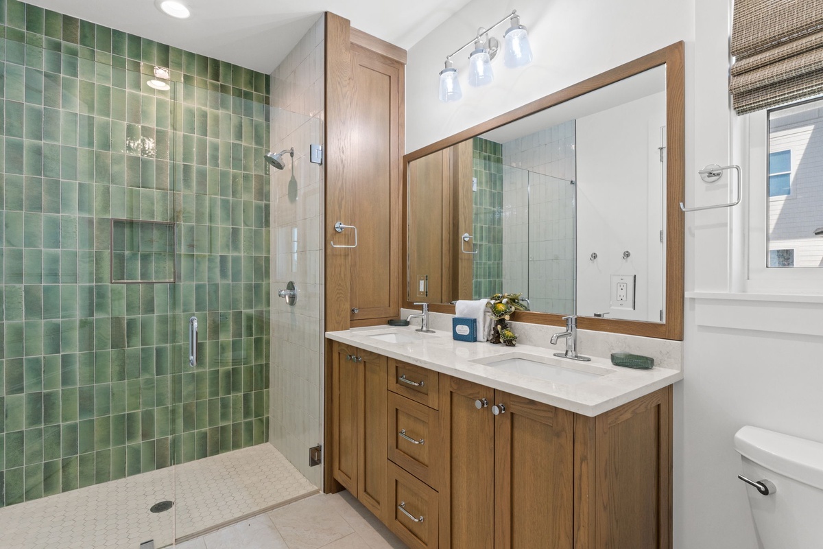 Step into your spa-like bathroom featuring a walk-in shower with calming green tile, double vanity with sleek white countertops, and natural light streaming through the window.