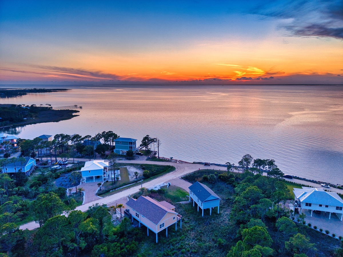 Aerial view of waterfront vacation homes nestled along a peaceful bay, with a stunning sunset painting the sky in brilliant shades of orange and gold.