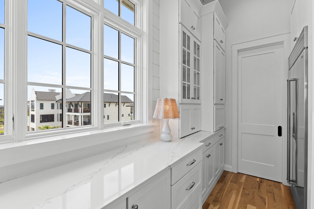 Your bright, airy kitchen features elegant white cabinetry, gleaming marble-style counters, and tall windows flooding the space with natural light—ideal for preparing meals with a view.