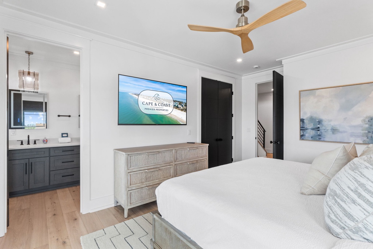 Your beautifully appointed bedroom features crisp white linens, elegant coastal-inspired furnishings, and a bright en-suite bath, all cooled by a modern ceiling fan for your total comfort.