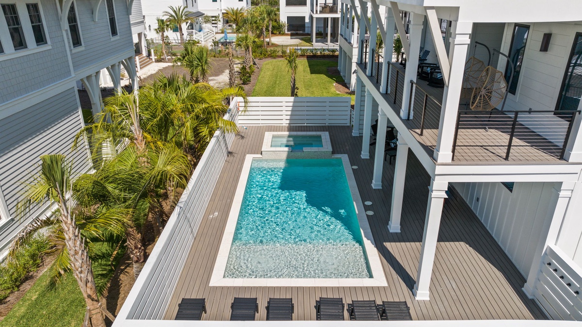 Aerial view of a modern coastal property featuring a private pool, hot tub, deck, and landscaped gardens surrounded by residential buildings.