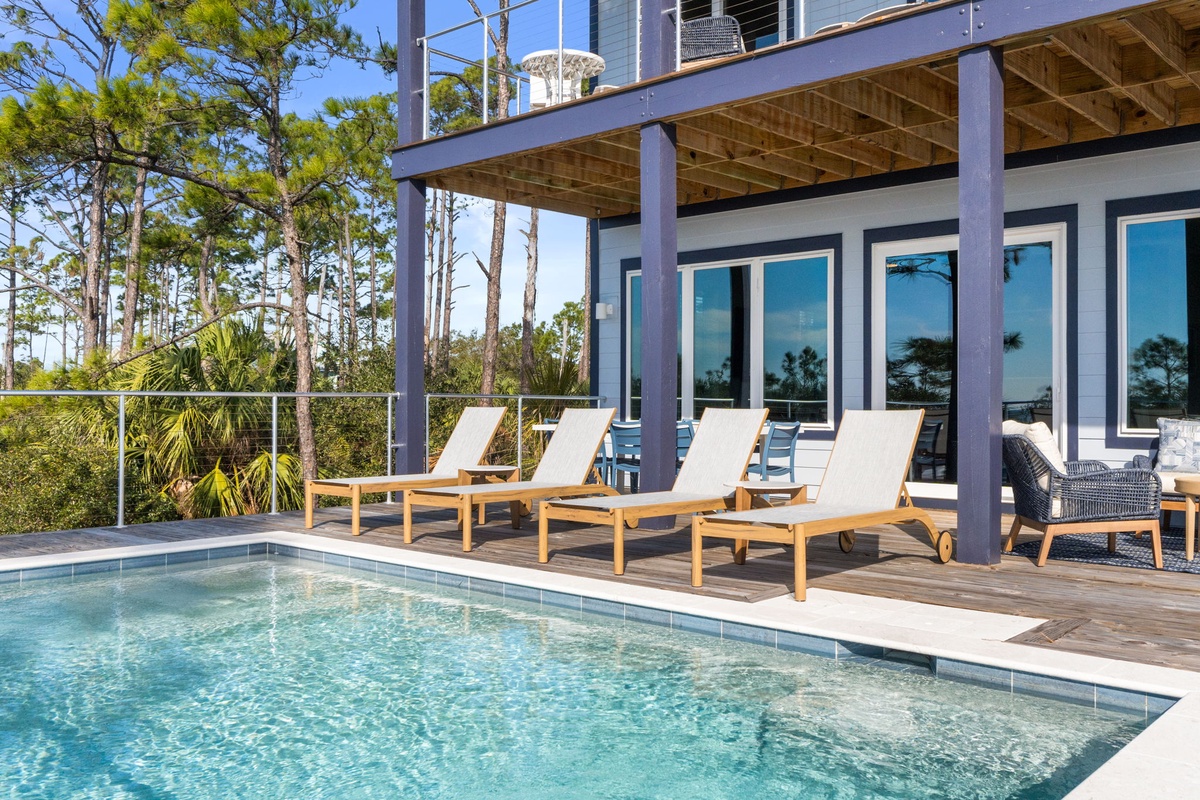 Lounge poolside on teak chaises under the covered deck, where floor-to-ceiling windows frame your private oasis amid lush coastal pines.