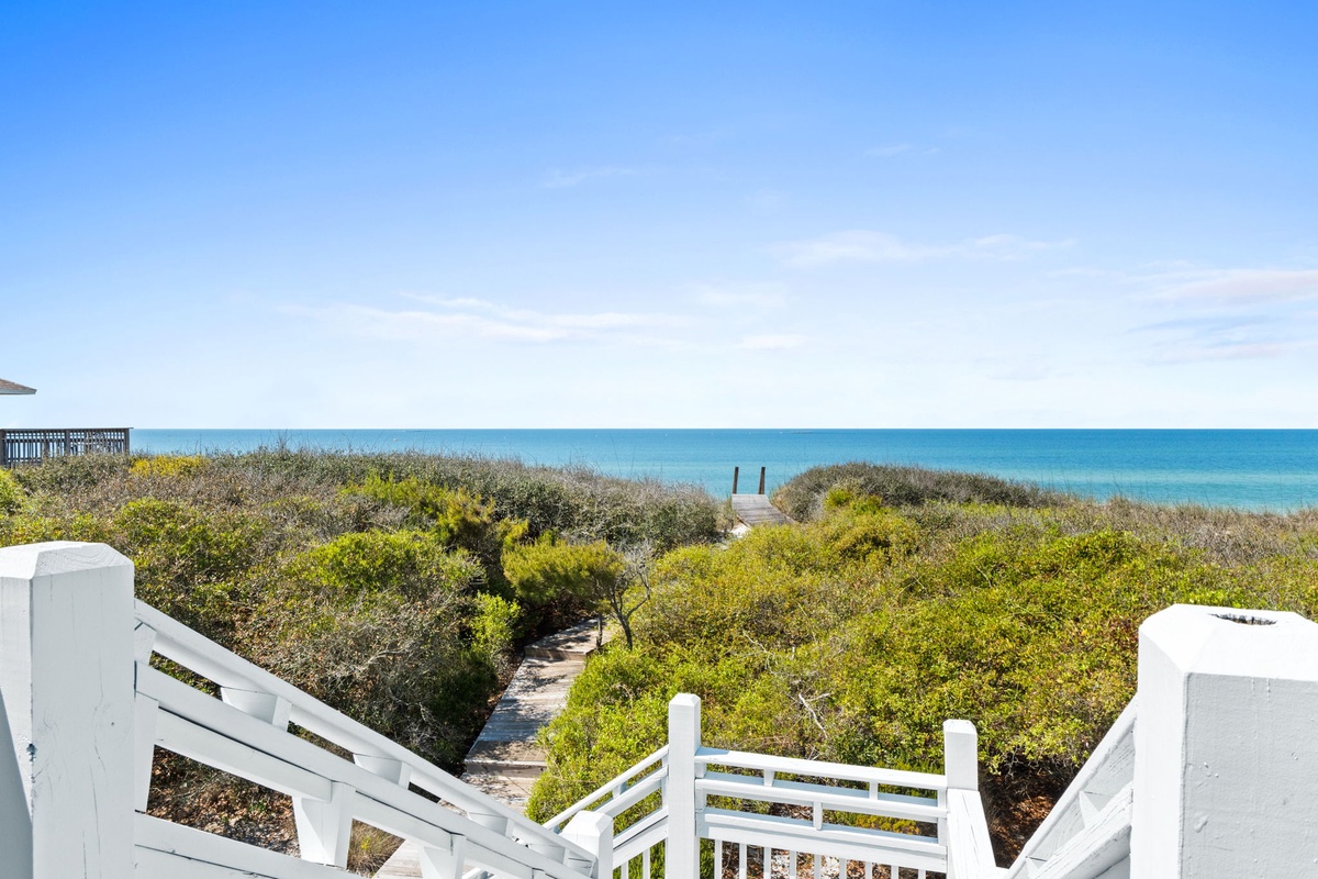 White wooden stairs lead down through coastal dunes to pristine turquoise waters. Natural vegetation frames the sandy pathway to the beach.