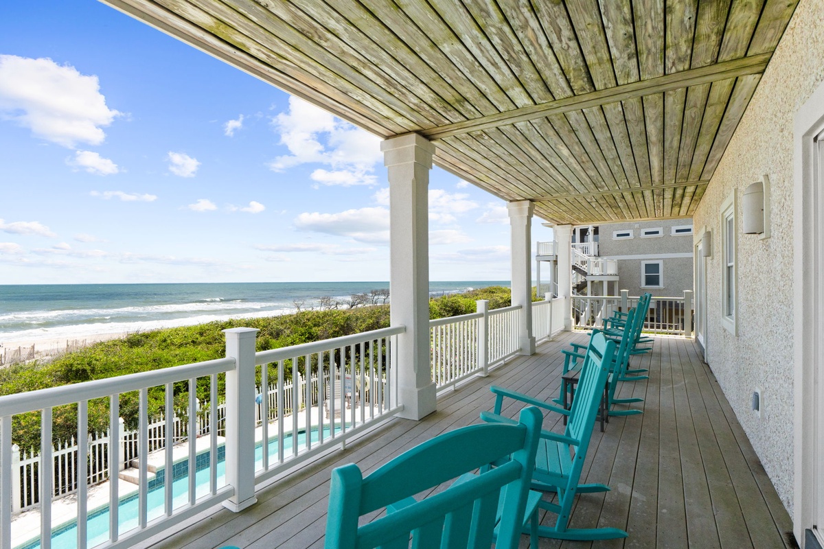 A covered beachfront deck featuring comfortable turquoise seating, offering a direct view of the ocean and coastal greenery. Part of a multi-level vacation property situated along the shoreline.