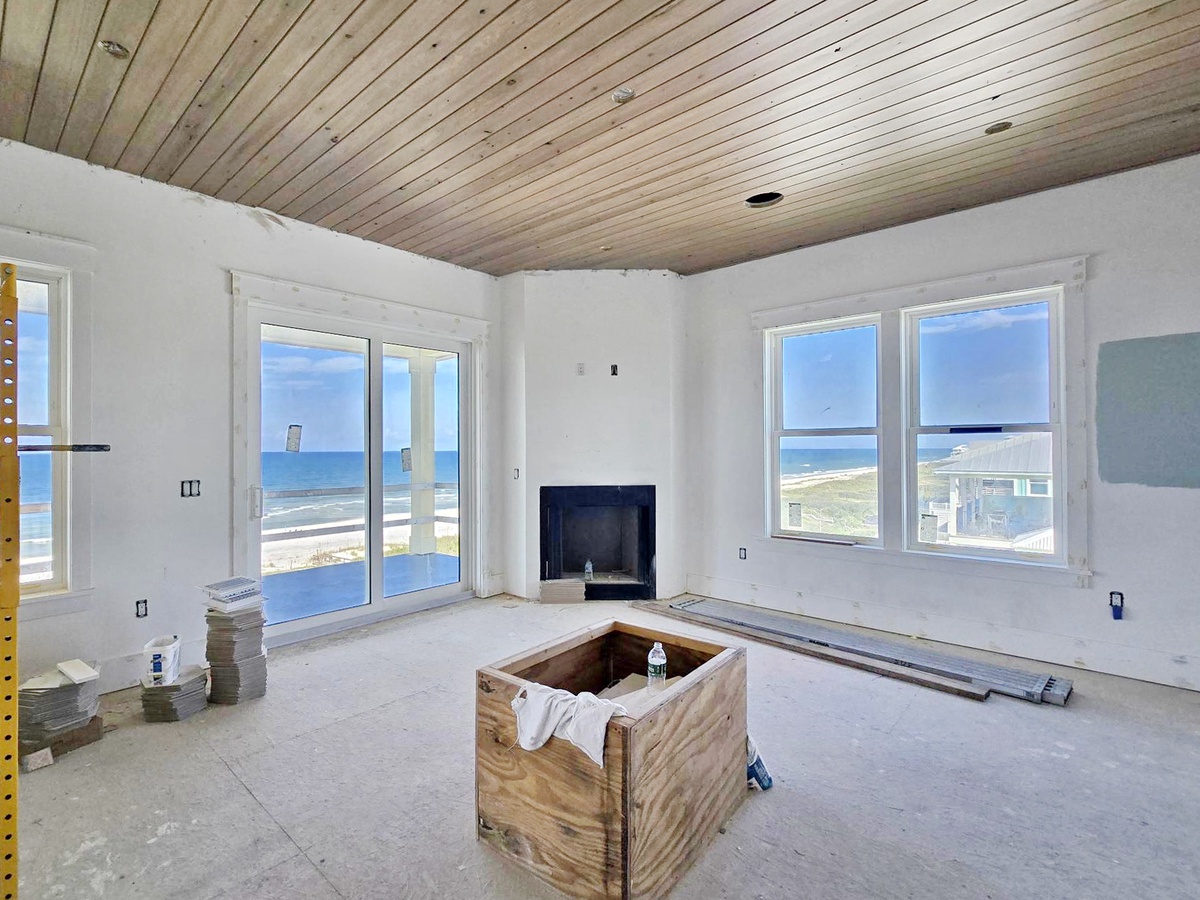 This oceanfront living space under renovation will soon offer stunning beach views through expansive windows and glass doors, with a warm wood-paneled ceiling adding coastal charm.