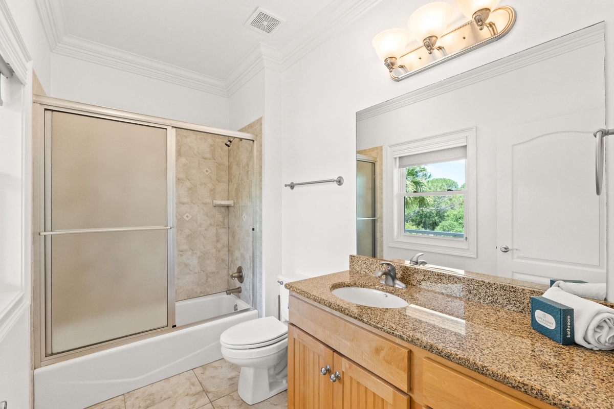 Your private bathroom features a granite vanity, glass-enclosed shower-tub combo, and a window overlooking natural greenery for a peaceful, refreshing retreat.