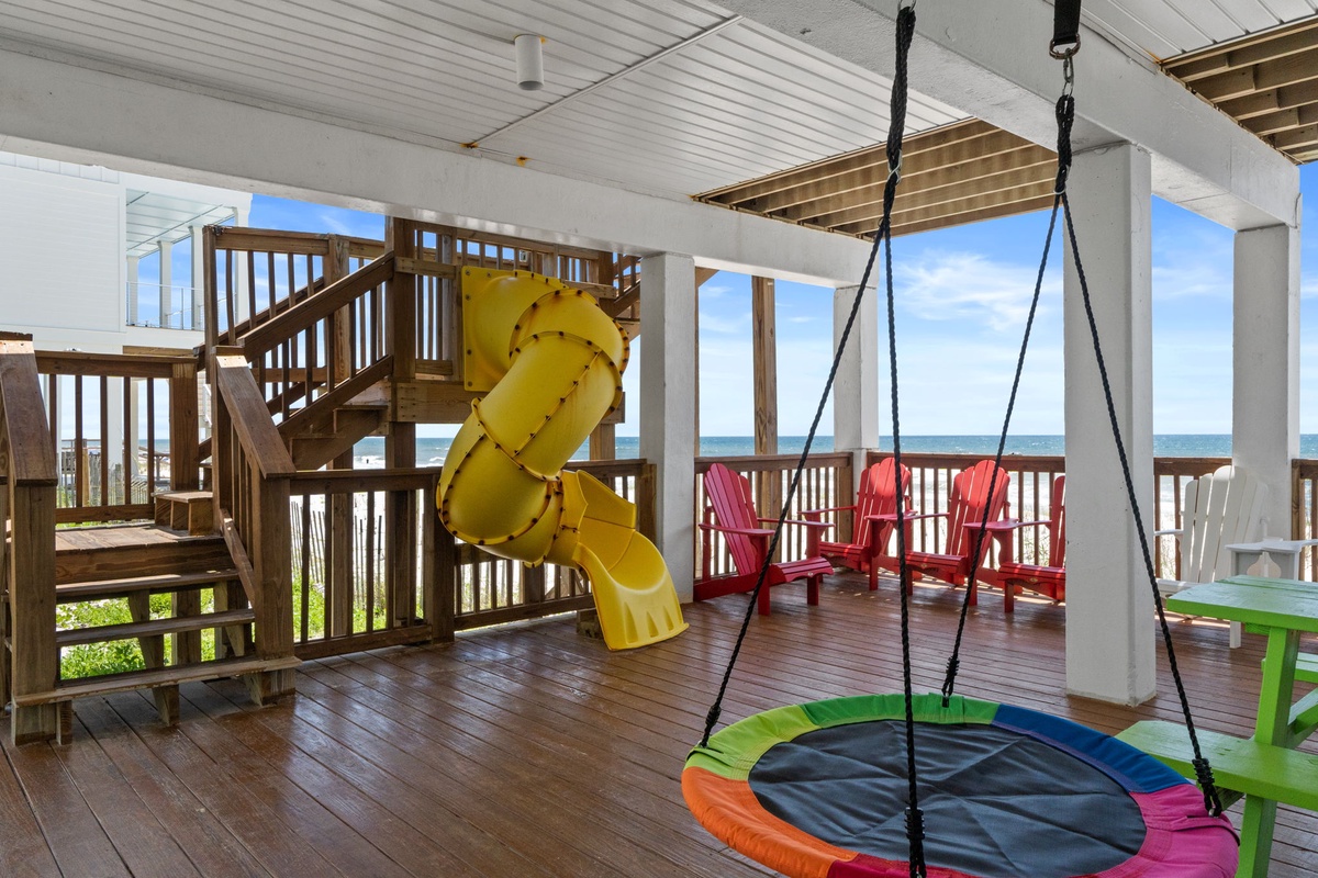 A covered deck overlooking the Gulf offers your family endless entertainment: a twisting yellow slide, a saucer swing, red Adirondack chairs, and stunning sea views perfect for unforgettable beach days.