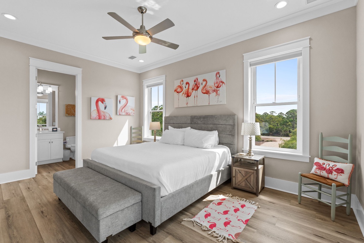 Your charming bedroom retreat features elegant flamingo artwork, wood floors, and windows revealing peaceful views of greenery and water. Ceiling fan and bright natural light create a breezy, coastal atmosphere.