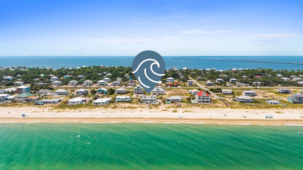 This beachfront community offers direct access to pristine white sands and calm turquoise waters, with vacation homes nestled along the shore and a distant bridge stretching across the bay.