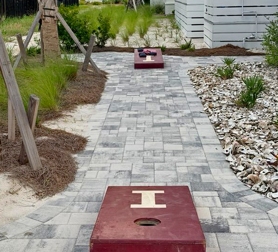 A paver walkway leads to outdoor cornhole boards set among landscaped gardens—perfect for friendly competition during your coastal getaway.