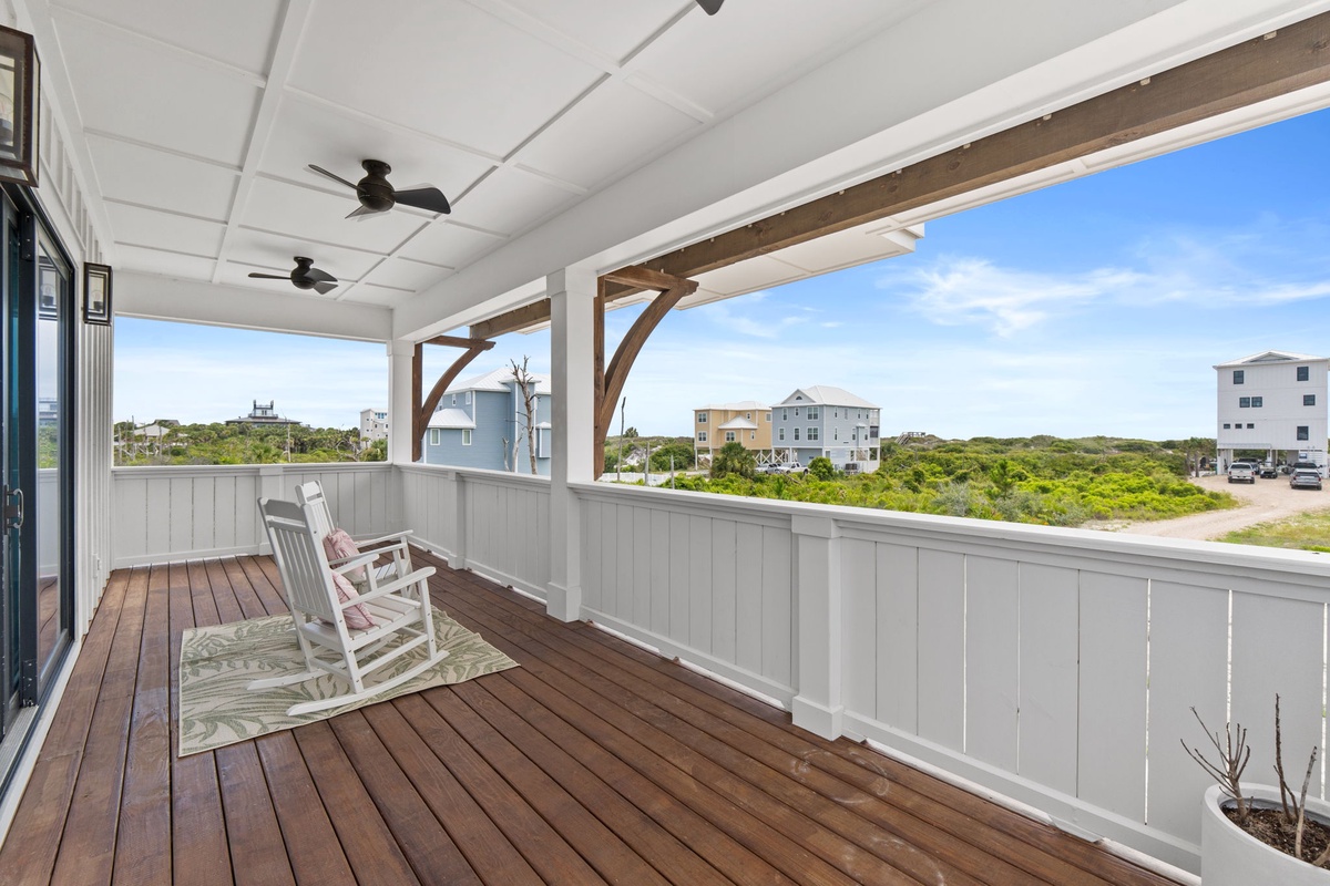 A covered porch with wooden decking and a rocking chair, overlooking nearby coastal buildings and lush greenery, perfect for morning coffee or evening relaxation.