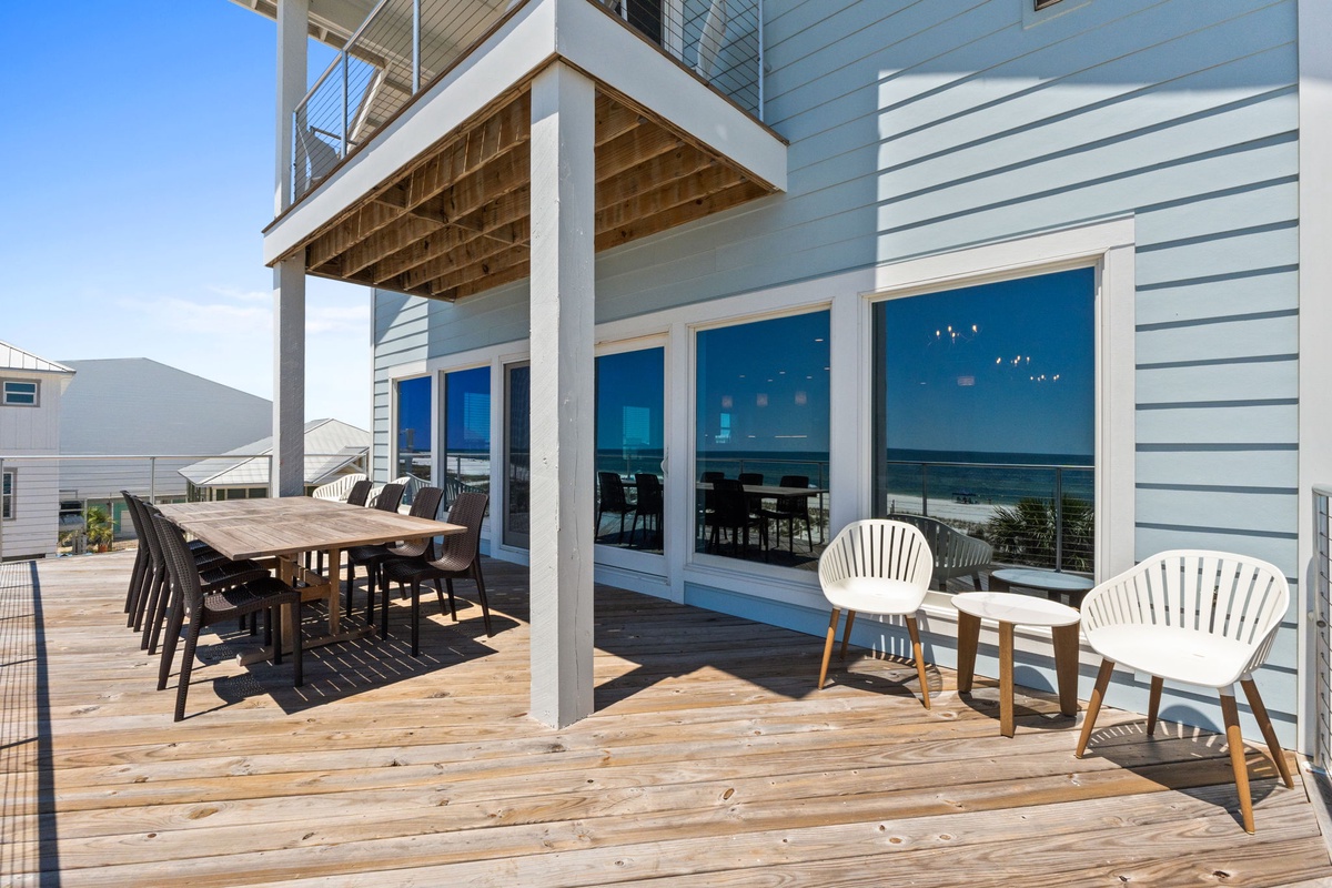 Your spacious deck features modern outdoor seating, a long dining table for group meals, and glass doors opening to a bright interior—all with stunning ocean views stretching to the horizon.