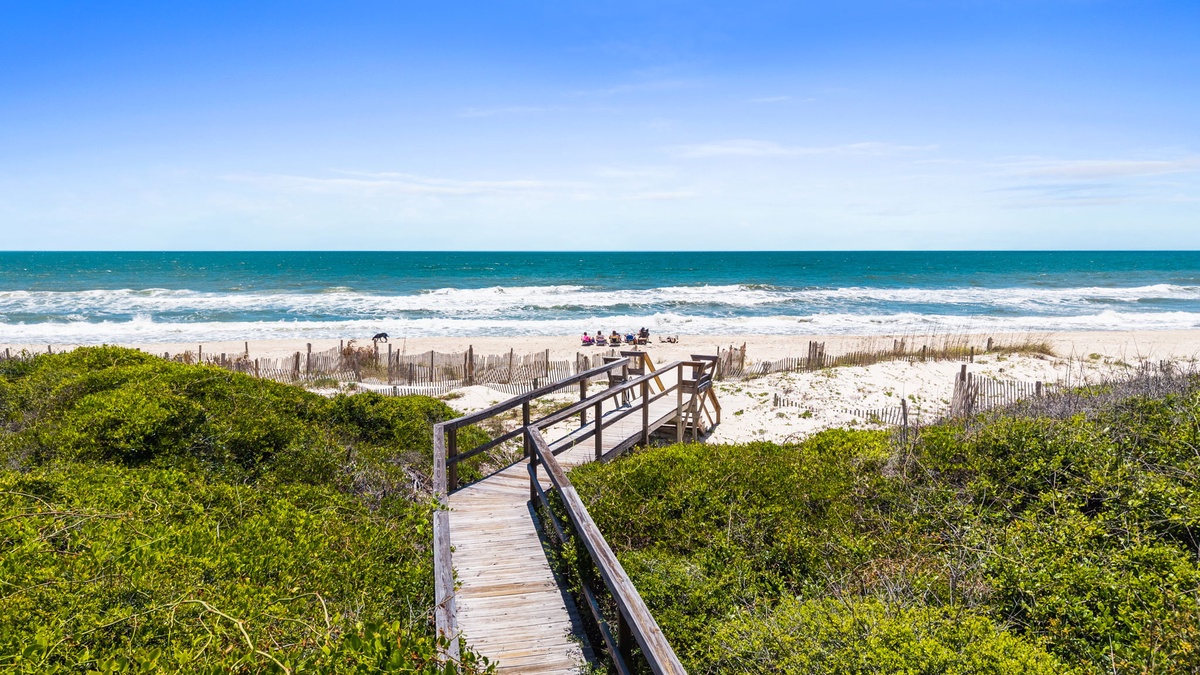 A charming wooden boardwalk winds through coastal dunes toward pristine white sands and turquoise waters, marking your direct beach access.