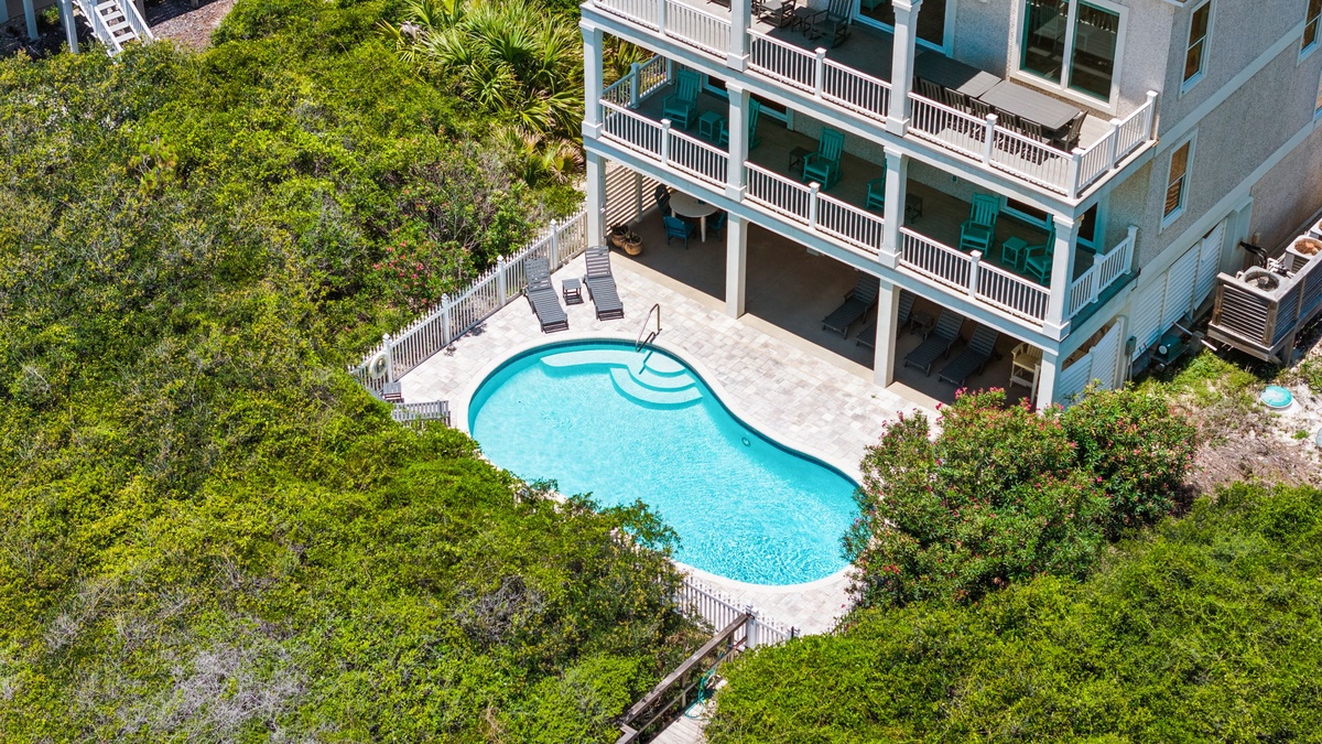 Aerial view of a coastal vacation rental featuring a private pool, multiple balconies with turquoise railings, and lush surrounding greenery near the beach.