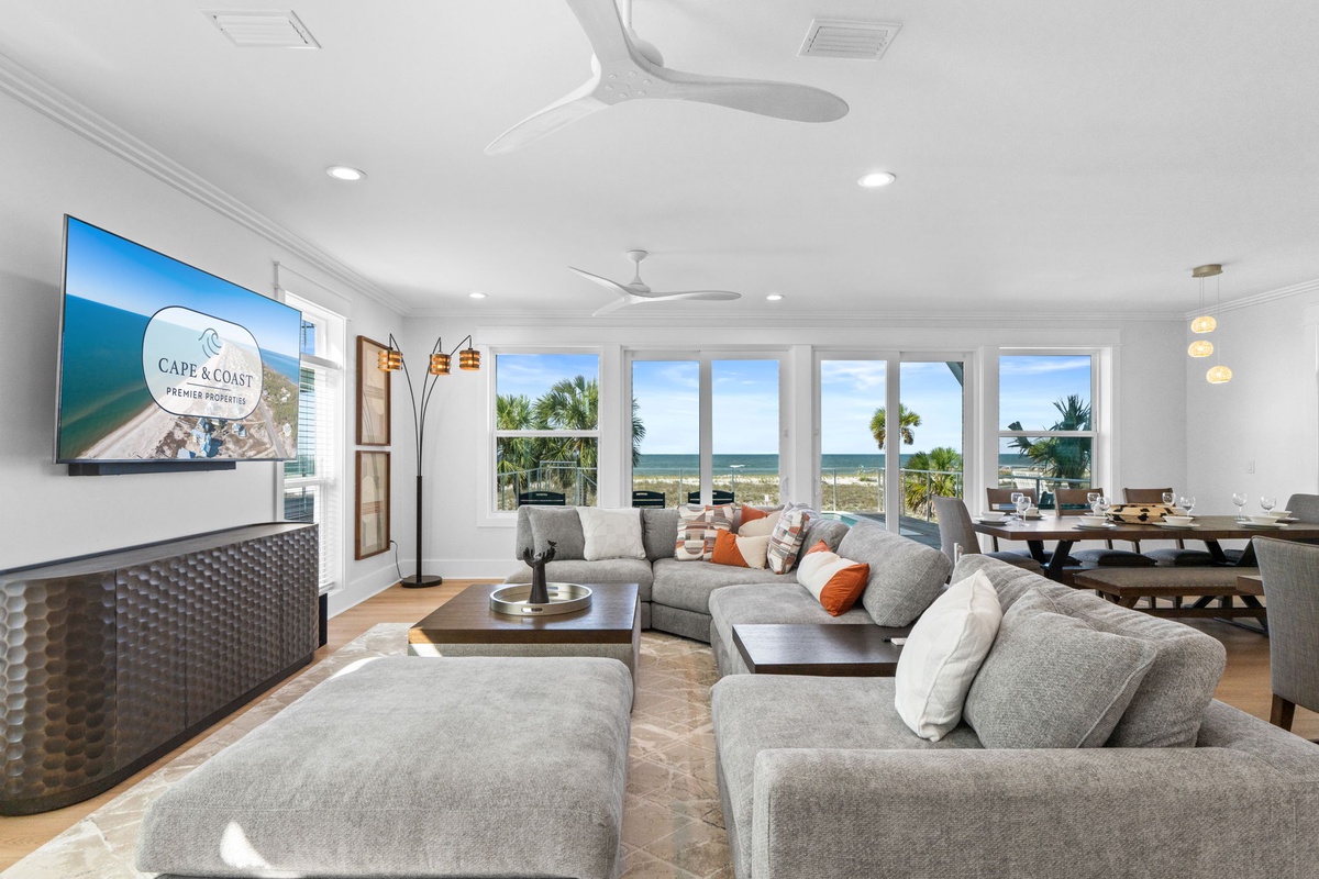 Your beachfront living room blends comfort and style, with plush seating, ceiling fans, and expansive windows framing endless ocean and palm tree views—perfect for unwinding in coastal elegance.