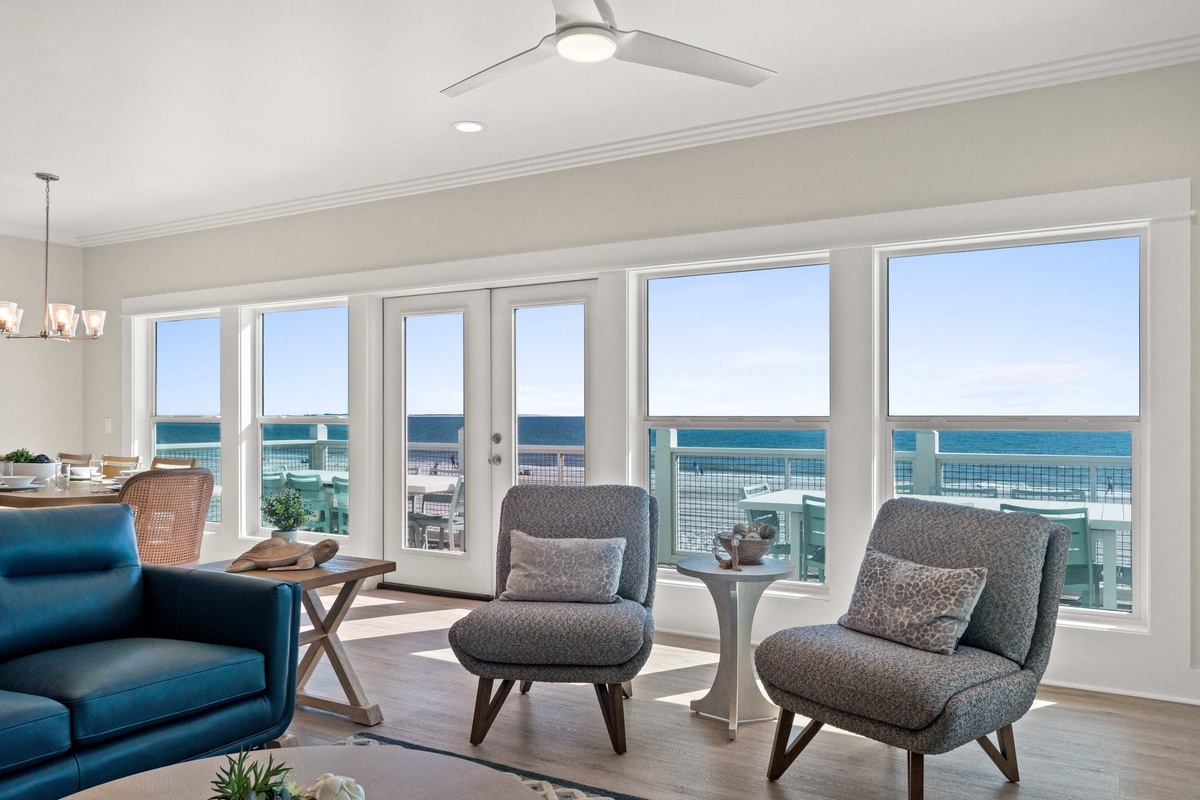 Relax in your bright living room with panoramic ocean views through floor-to-ceiling windows, complete with comfortable seating and coastal breezes from the ceiling fan.