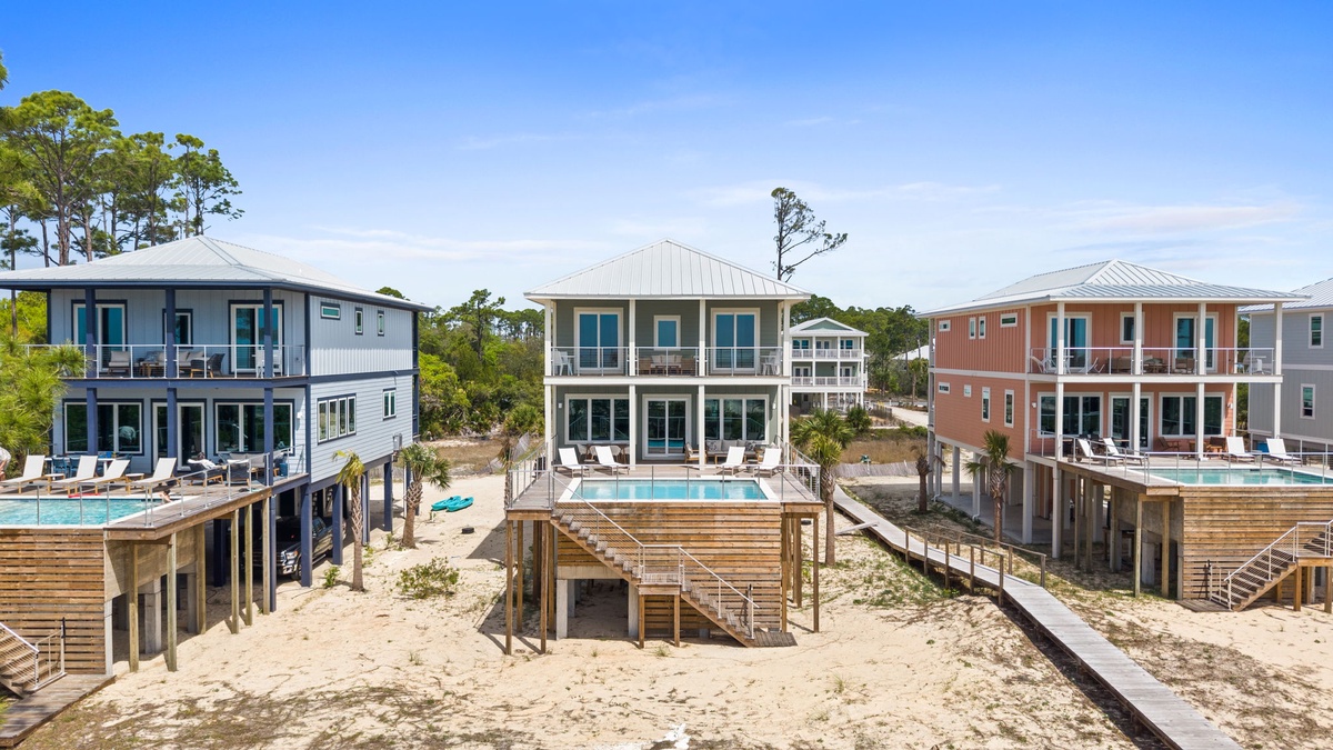 Modern beachfront vacation homes with elevated pools, wooden walkways, and colorful coastal architecture nestled among natural dunes and trees.