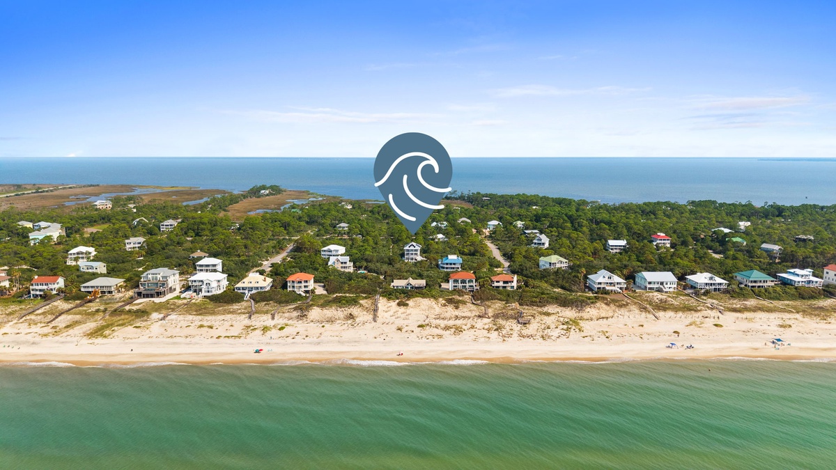 Aerial view of a serene coastal neighborhood, with beach homes tucked among trees along a pristine sandy shore and calm turquoise waters.