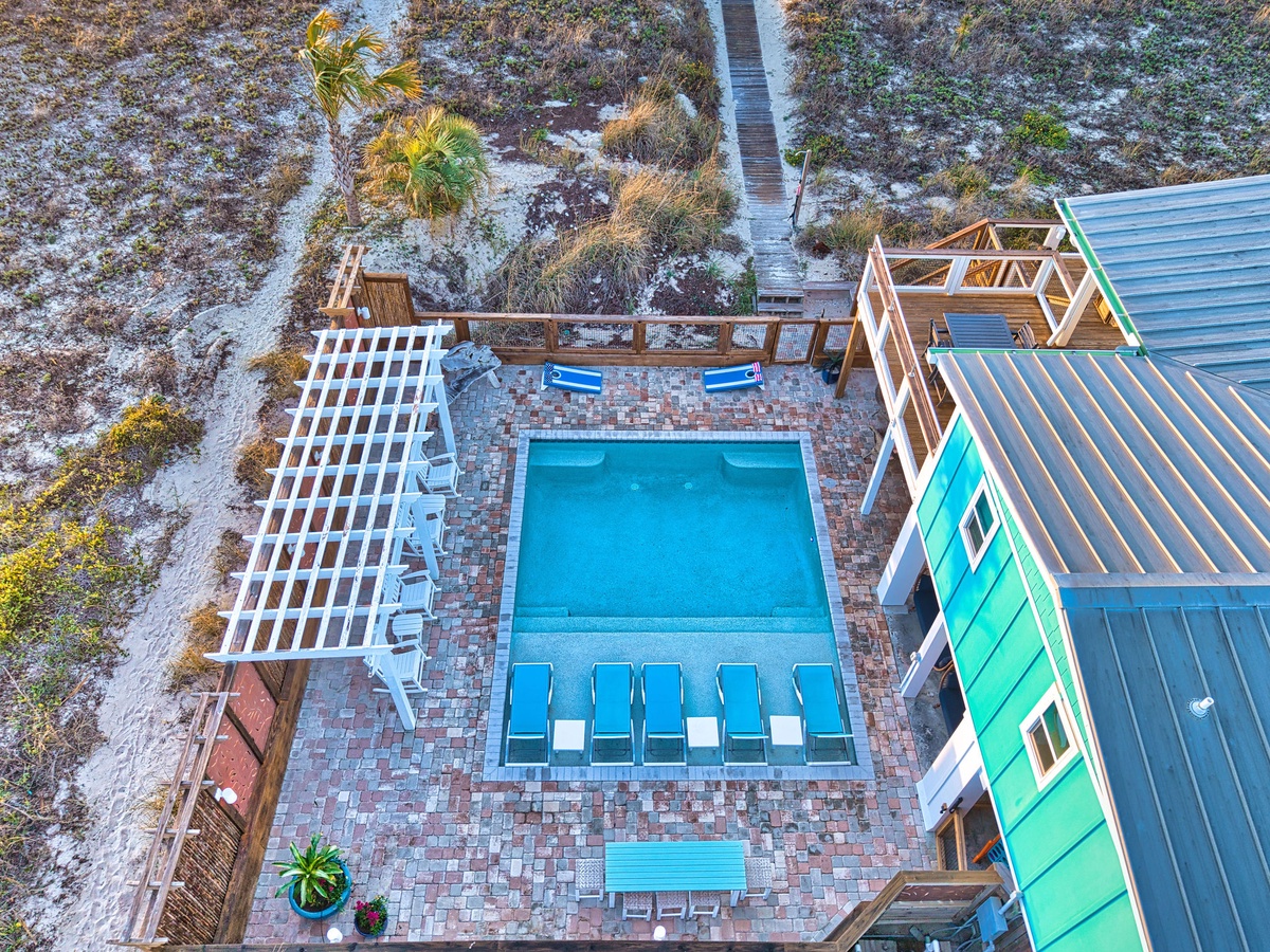 Aerial view of a coastal vacation rental featuring a stunning swimming pool surrounded by brick patio and natural dune landscape.