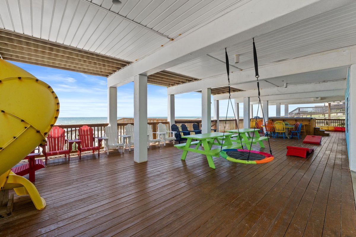 Covered oceanfront deck features colorful Adirondack chairs, a swing table, and cheerful play equipment—your family's perfect spot to relax while the waves roll in.