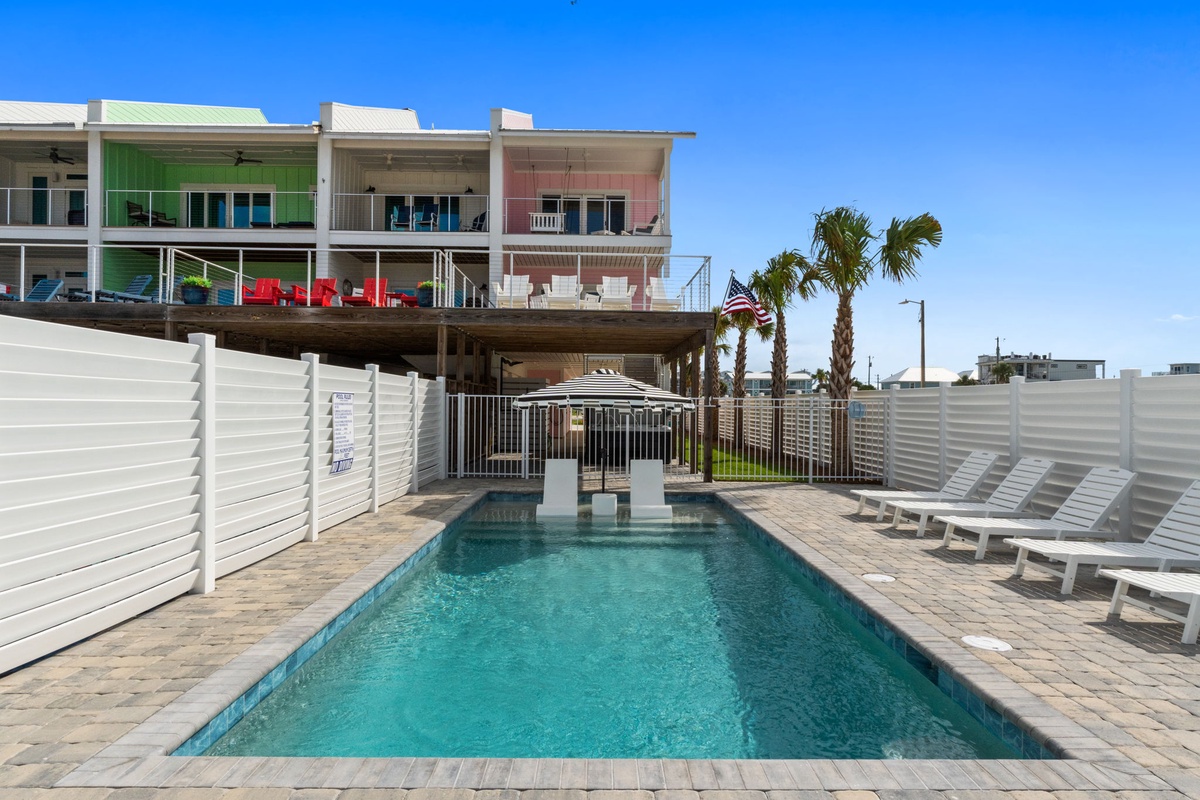 A sparkling pool surrounded by loungers awaits beneath palm trees and blue skies, with colorful balconies overlooking the inviting outdoor space.