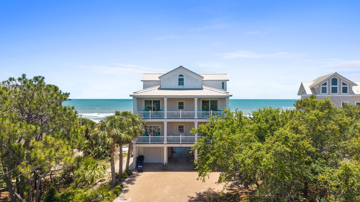 Coastal beachfront property featuring a multi-story home with double-level porches overlooking turquoise waters, nestled among palms and native vegetation with direct beach access.