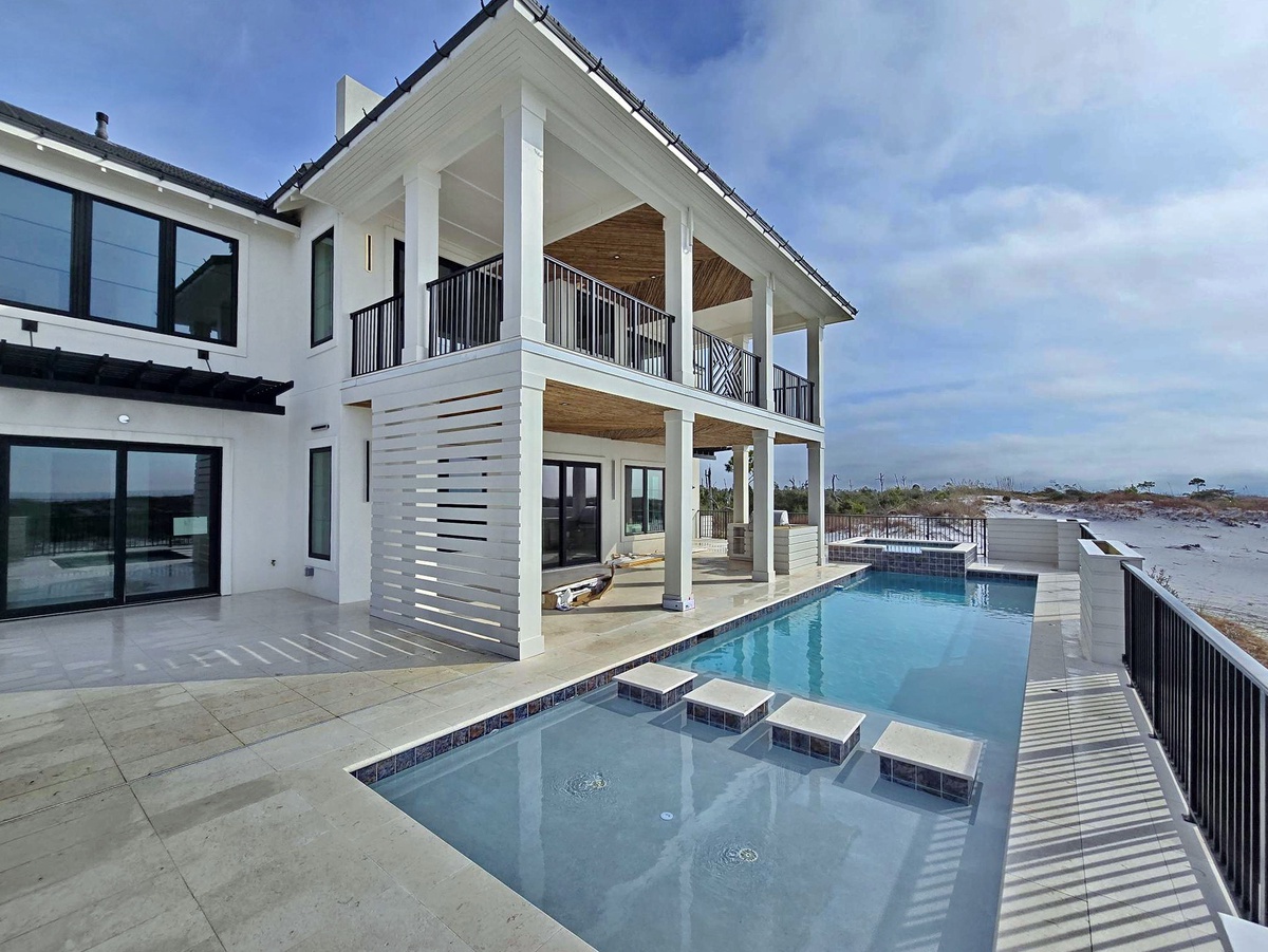 Modern coastal luxury home featuring elegant white columns, expansive covered terraces, and pristine swimming pool overlooking peaceful waterfront surroundings.