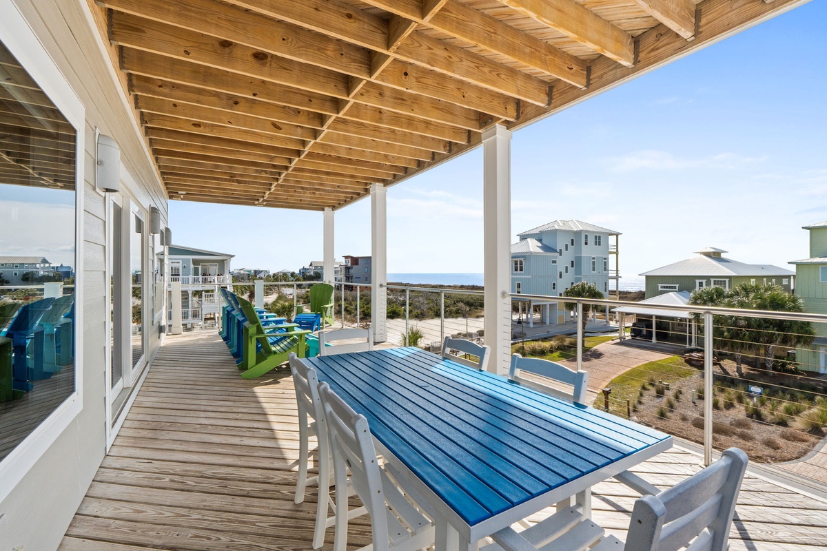 Your spacious deck invites coastal dining under protective beams, with colorful chairs and ocean glimpses beyond.