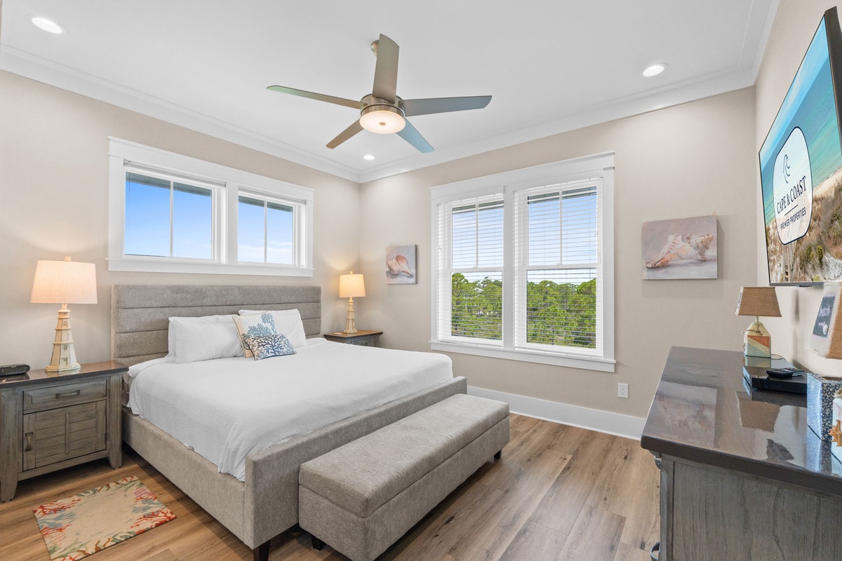 Step into this serene bedroom where natural light and breezy coastal touches create your perfect retreat. You'll love waking up to views of surrounding greenery through large windows while a ceiling fan keeps things comfortable year-round.