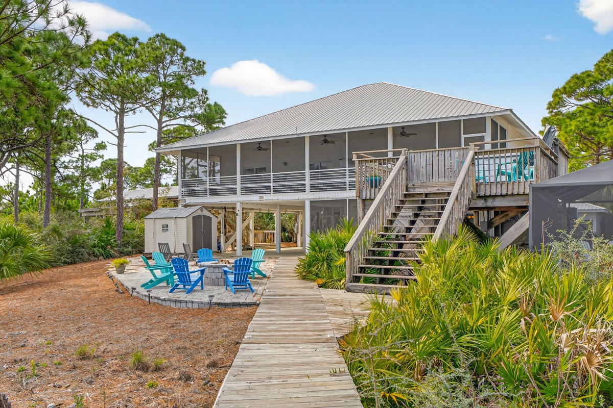 Elevated beach house with wraparound porch and colorful Adirondack chairs surrounding a fire pit, nestled among coastal pines.