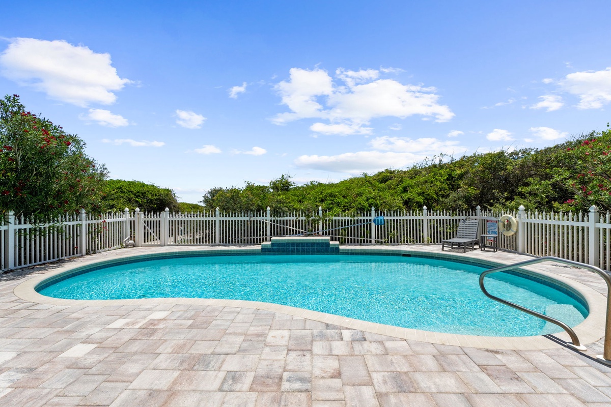A spacious swimming pool framed by lush greenery, featuring a curved design and paver deck under clear blue skies. Perfect for sunny afternoon relaxation.