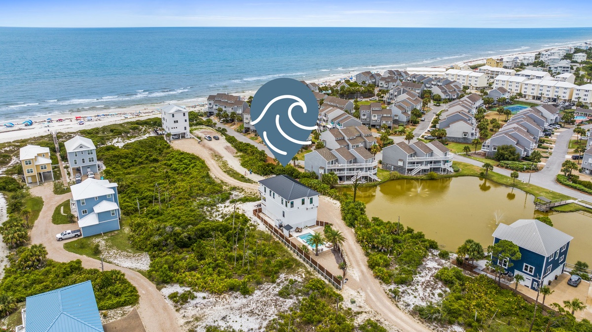 Aerial view of a beachside vacation rental community with colorful homes nestled among palms and dunes, steps from the sandy shoreline and ocean waves.