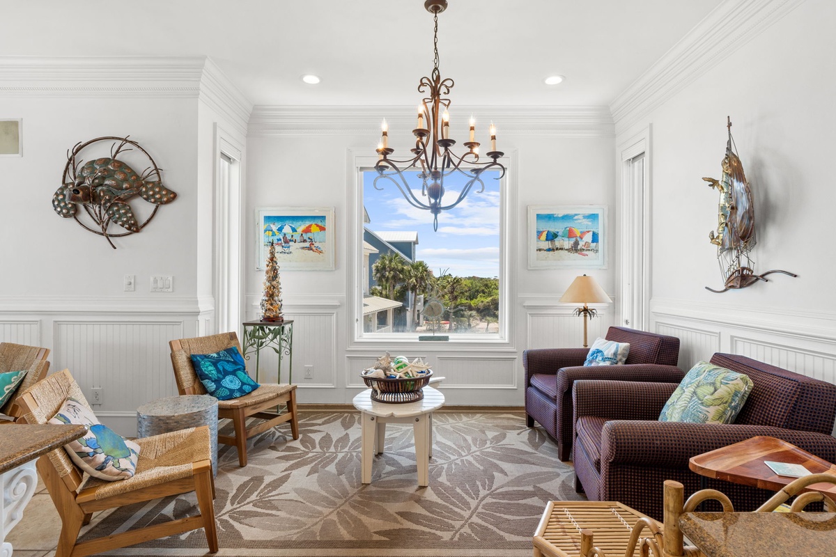 Relax in your coastal-inspired living room with beach-themed art and comfortable seating, framed by bright natural light from the window overlooking lush greenery.