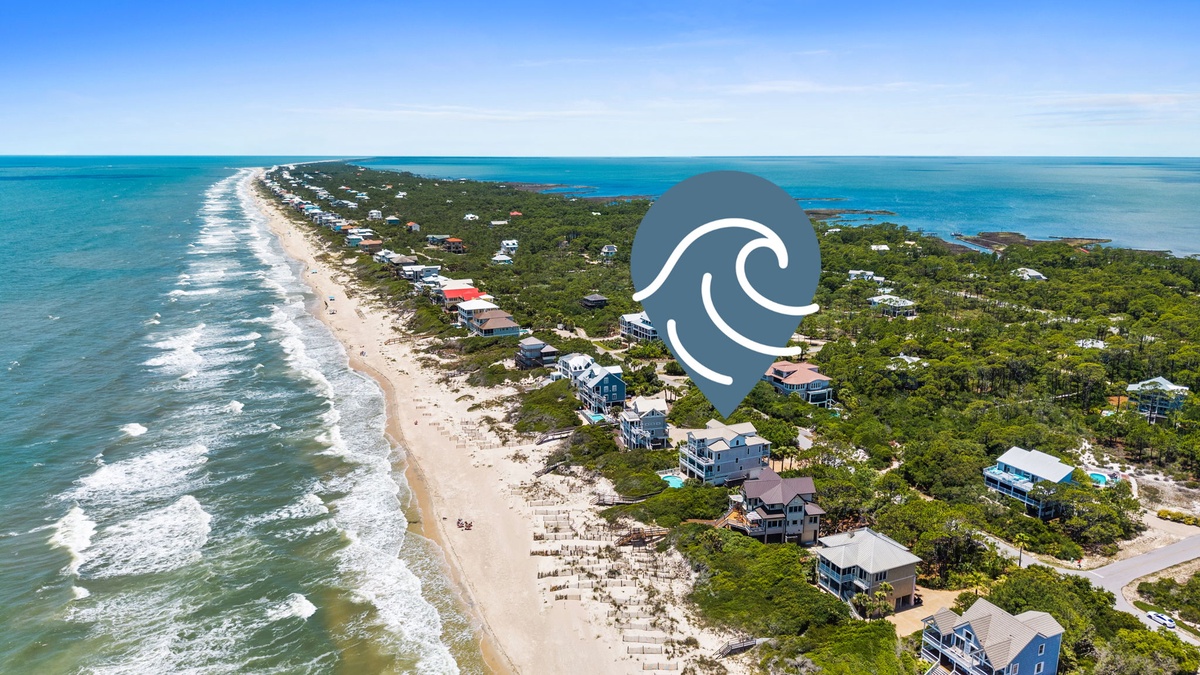 An aerial view captures a stunning beachfront community nestled between pristine ocean waters and lush coastal vegetation, showcasing the property's idyllic island setting.