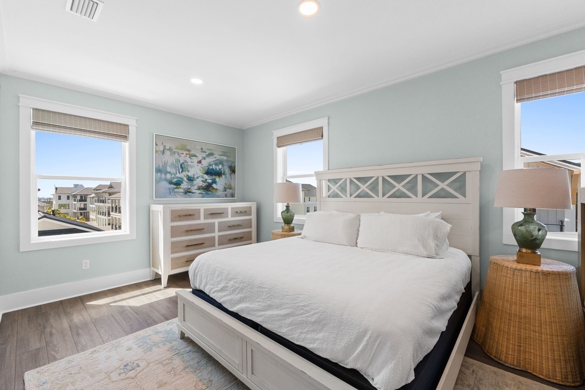 Drift off in this serene coastal bedroom, where soft seafoam walls and crisp linens invite total relaxation. Natural light pours through three windows, framing glimpses of the neighborhood and sky beyond.