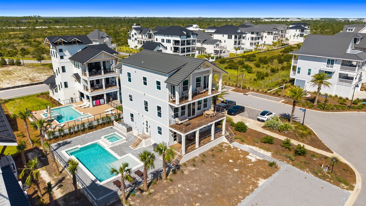 An aerial view of a coastal neighborhood featuring modern vacation homes, private pools, and palm-lined streets amidst natural dune vegetation.