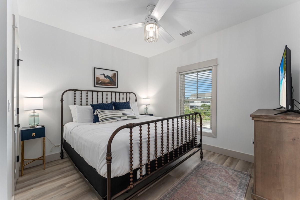 Rest peacefully in your stylish bedroom featuring a charming wrought iron bed, coastal artwork, and modern ceiling fan for ultimate comfort.