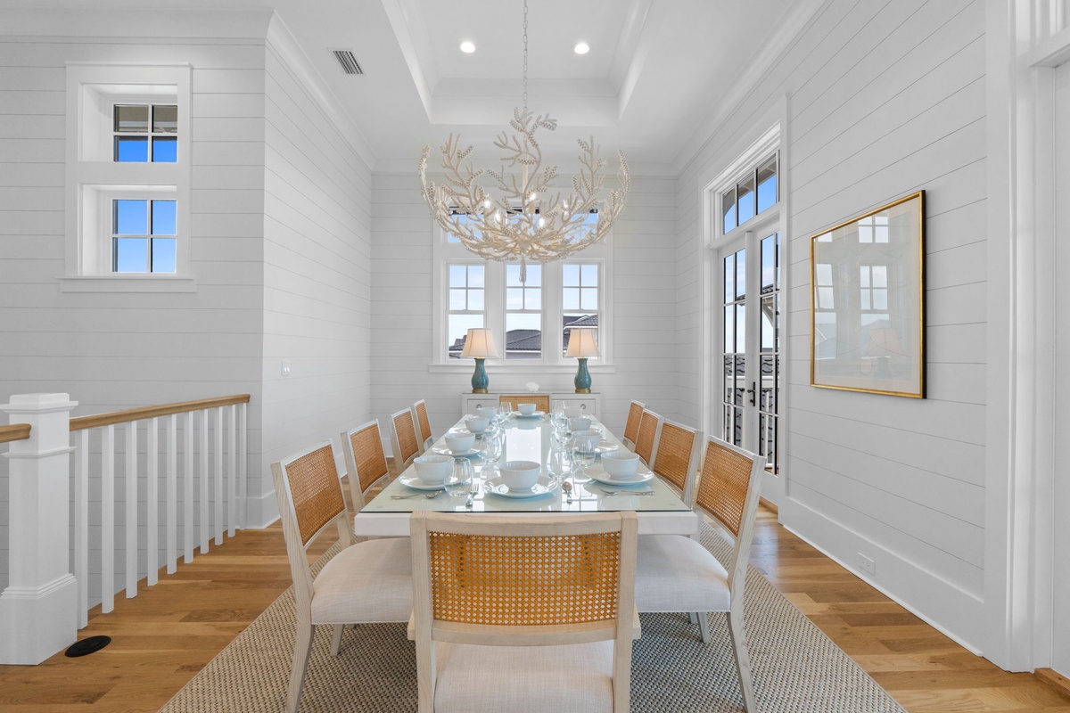Gather around this elegant glass-top dining table beneath a stunning coral chandelier, where natural light pours through tall windows and coastal-chic decor creates the perfect backdrop for your memorable meals together.