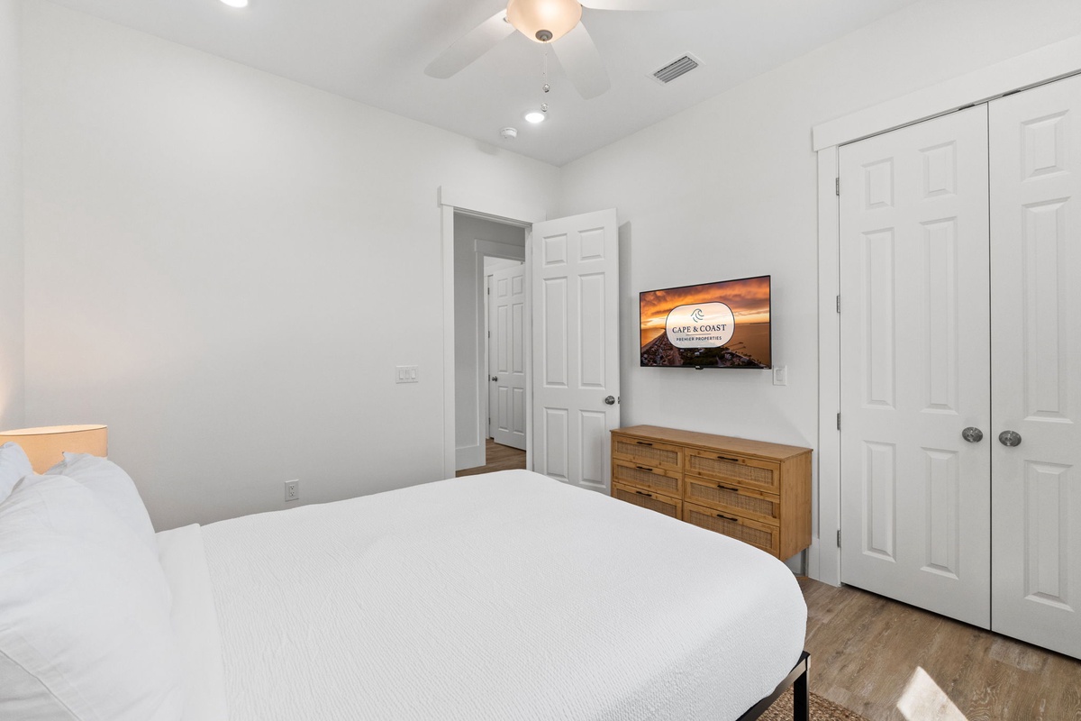 Unwind in your serene bedroom featuring crisp white linens, warm wood floors, and convenient entertainment with wall-mounted TV and spacious closet storage.