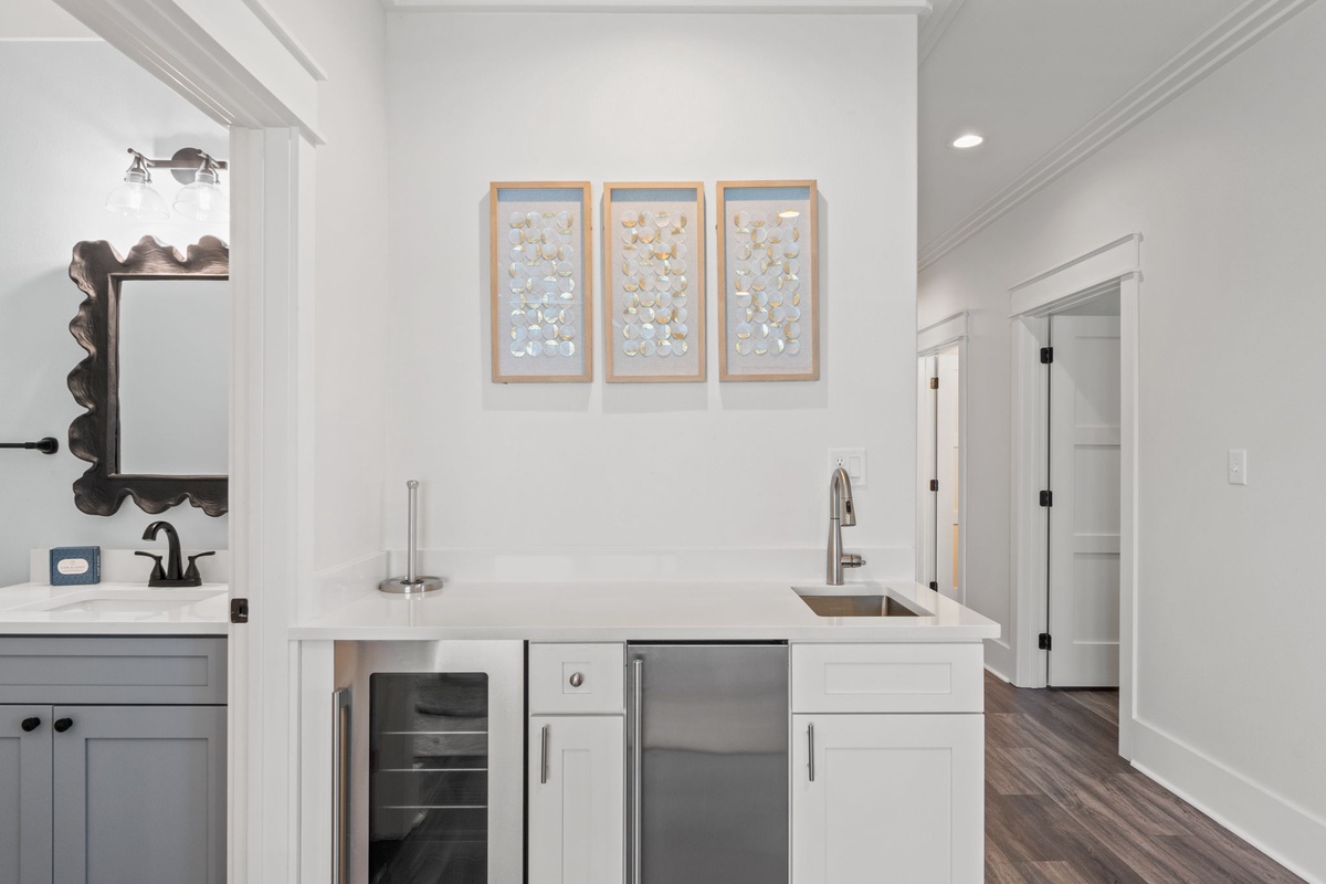 Your private wet bar offers everything you need to unwind—a sleek sink, wine cooler, and mini fridge, all framed by elegant coastal decor and warm lighting.