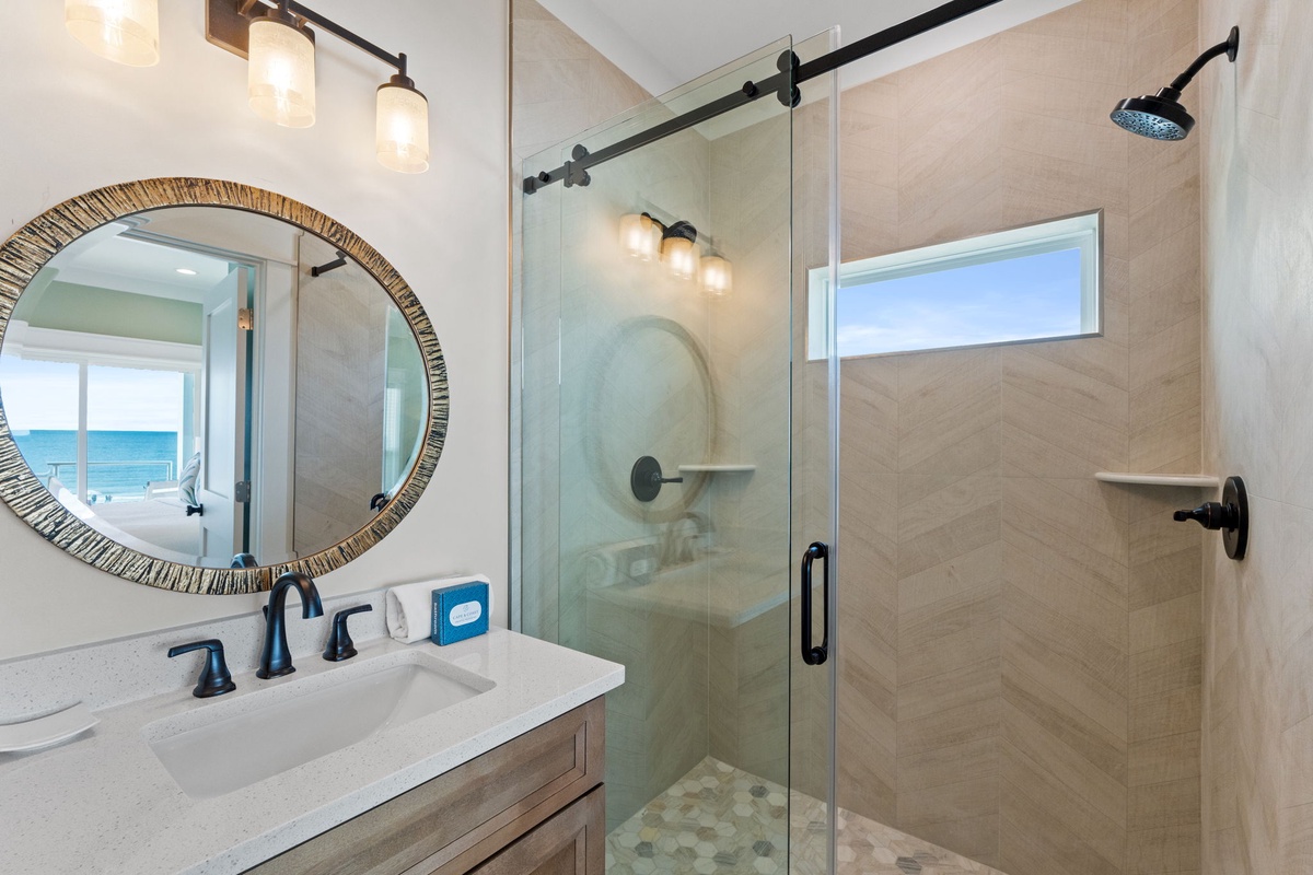 Your private bathroom features a spa-like walk-in shower with sleek glass doors and modern fixtures. Natural light fills the space, while thoughtful touches like the decorative mirror add warmth.